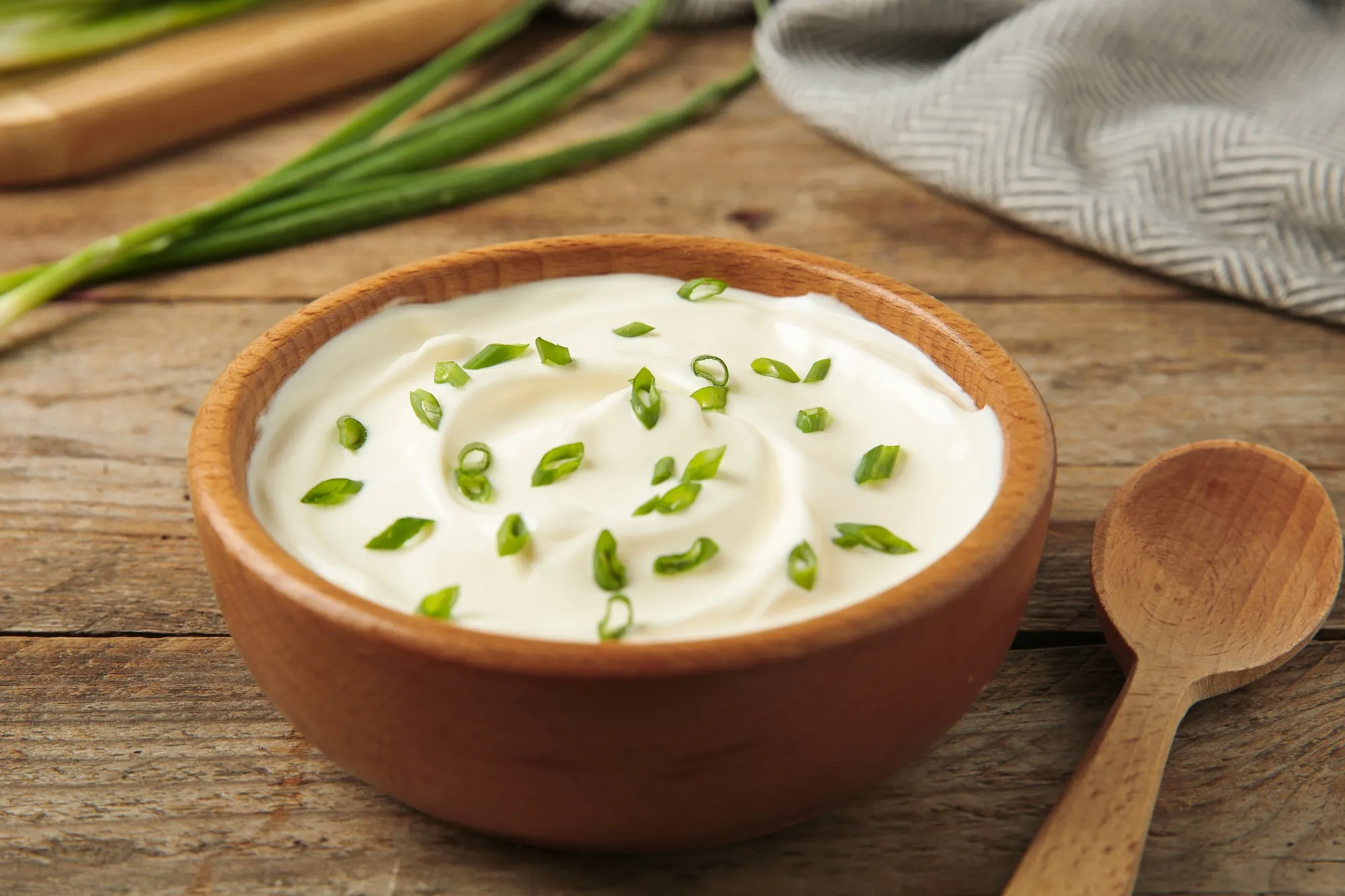 A wooden bowl of fresh sour cream topped with chopped green onions beside a wooden spoon on a rustic table